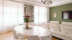 Ein zeitgemäßer Essbereich mit einem runden Tisch, weißen Stühlen, einer Vase mit Blumen, einem großen abstrakten Gemälde, einer Couch und einem Flachbildfernseher an der Wand.