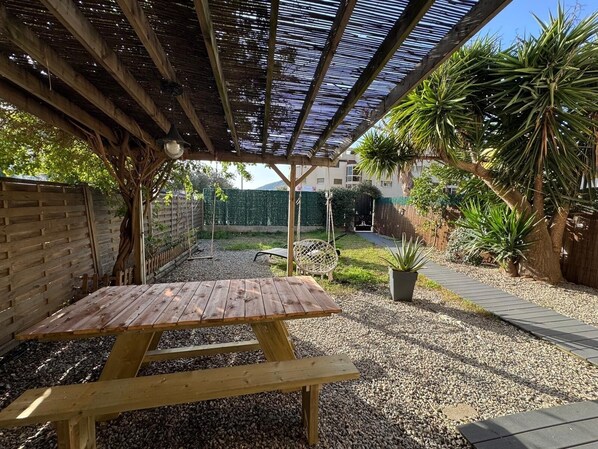 Una mesa de picnic de madera bajo una pérgola, con un camino de grava y plantas en macetas.