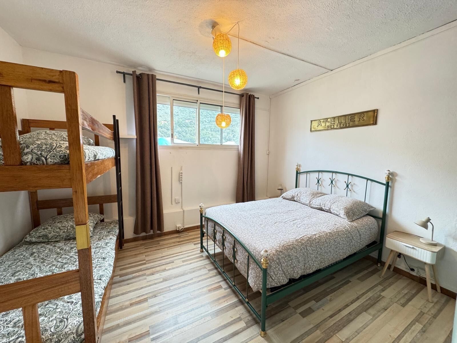 A room with a bunk bed, a single bed, a window with curtains, a ceiling light, and a small bedside table.