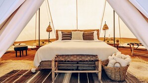 A tented accommodation with a bed, pillows, a bench, a basket, and a table with a lamp. - Magic Camps Wahiba Sands (Bidiya)