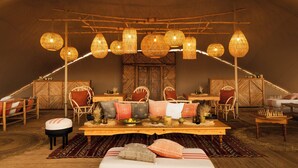 A tented room with wicker hanging lamps, wooden furniture, and a central table with a bowl of fruit. - Magic Camps Wahiba Sands (Bidiya)