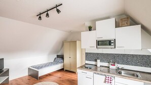 Et kompakt køkken med mikrobølgeovn, vask og skabe. Der er en bænk med et mønstret sædehynde og en lille plante oven på et skab.
