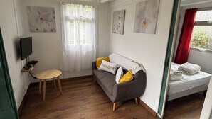 Een kamer met een grijze bank, een klein houten tafeltje, een televisie op een plank en een bed met een rood gordijn.