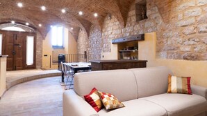 A rustic interior with stone walls, a wooden door, a dining area, and a sofa with a cushion. - Charming brick and stone loft in a 17th-century chapel in the historic center (Spello)