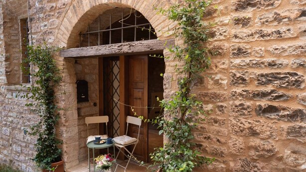 A stone building with an arched doorway, a small table with chairs, and potted plants. - Charming brick and stone loft in a 17th-century chapel in the historic center (Spello)