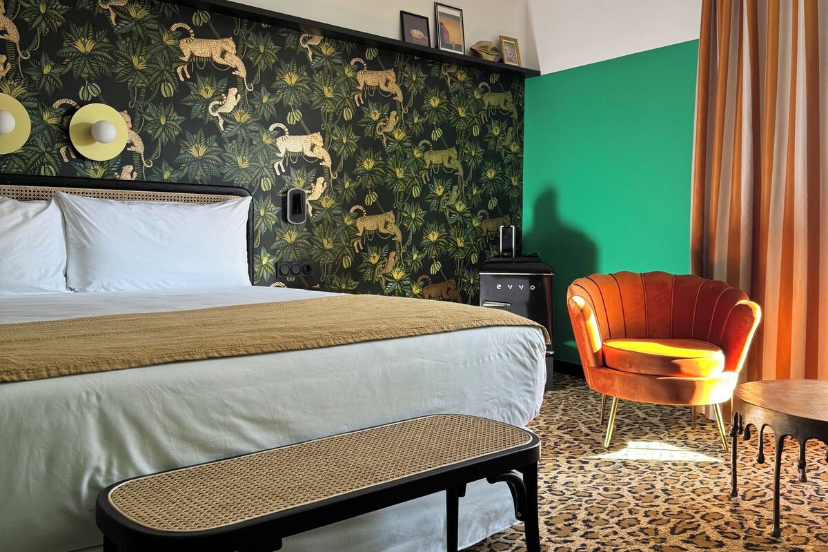 A hotel room with a bed, a bench, a chair, a desk, and a green wall with a jungle-themed wallpaper.