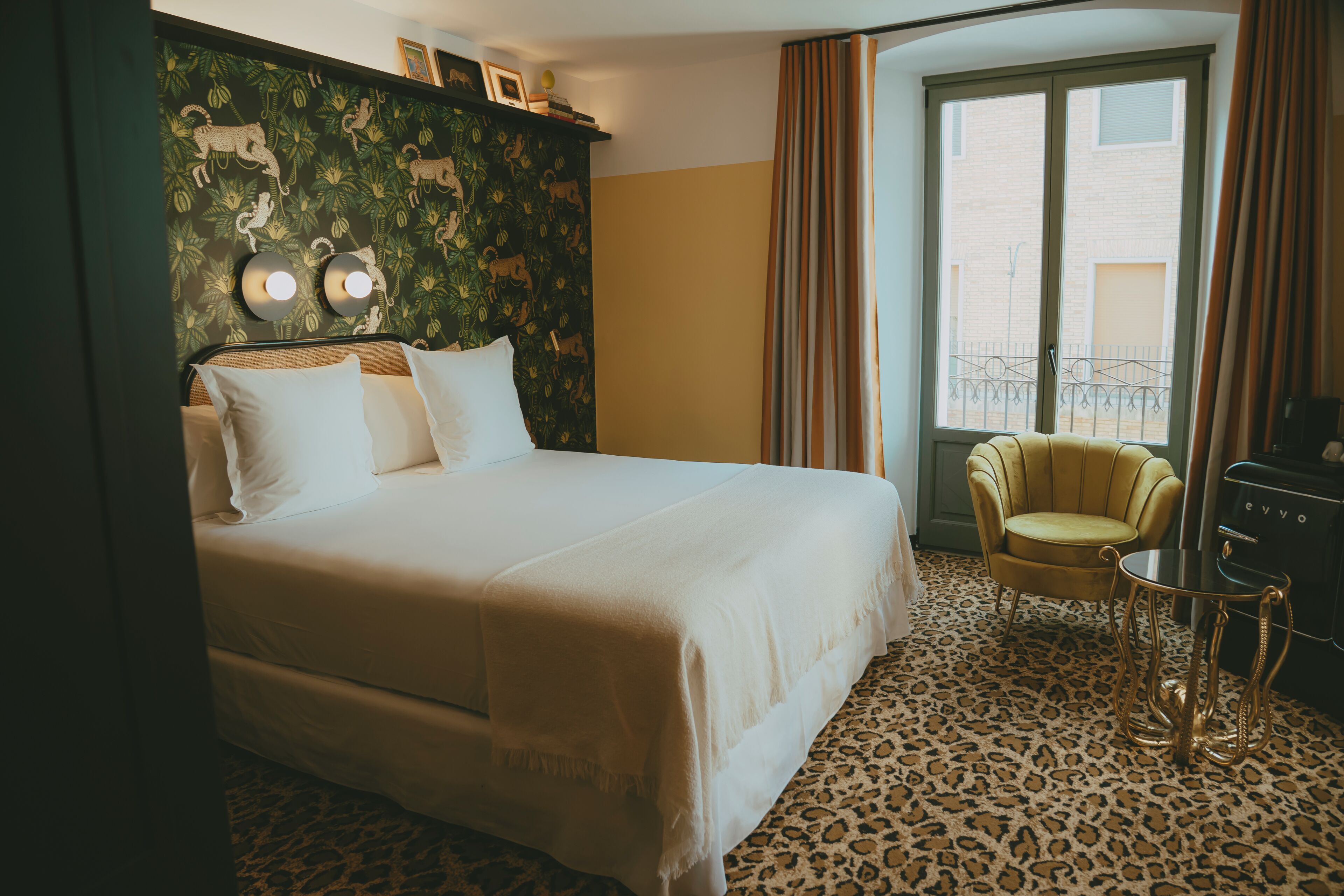 A hotel room with a bed, a chair, a table, a window with curtains, and a leopard print carpet.
