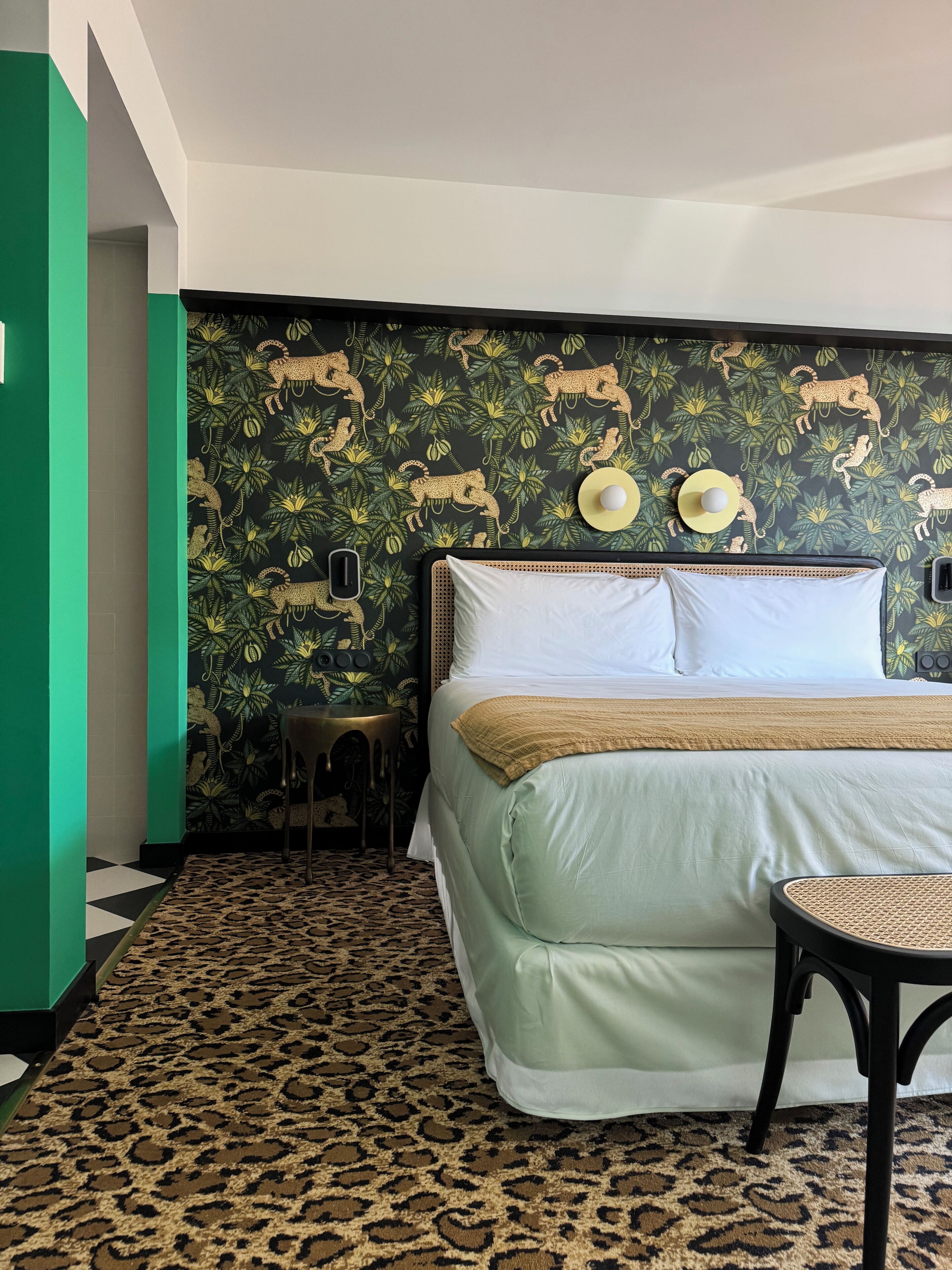 A hotel room with a bed, a small table, a chair, a patterned carpet, and a wall with a jungle-themed wallpaper.
