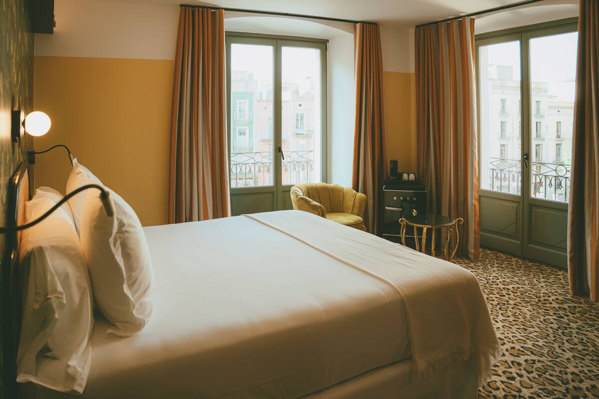 A hotel room with a bed, a chair, a desk, and a view of buildings through large windows.