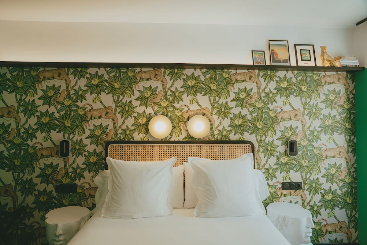 A bedroom with a bed, two bedside lamps, a shelf with pictures, and a wall with a jungle-themed wallpaper.