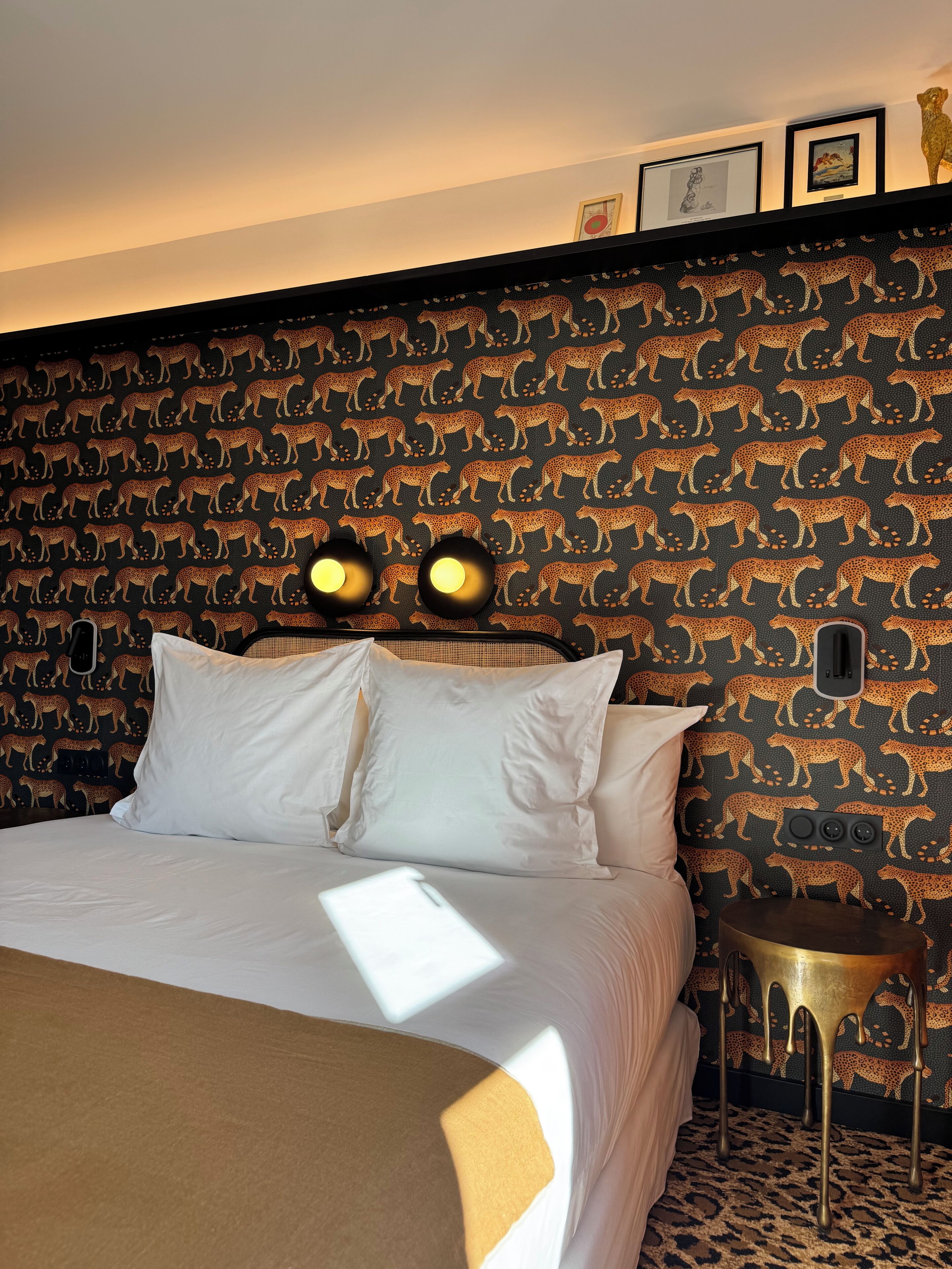 A hotel room with a patterned wallpaper, a bed with white linens, and a bedside table with a lamp.