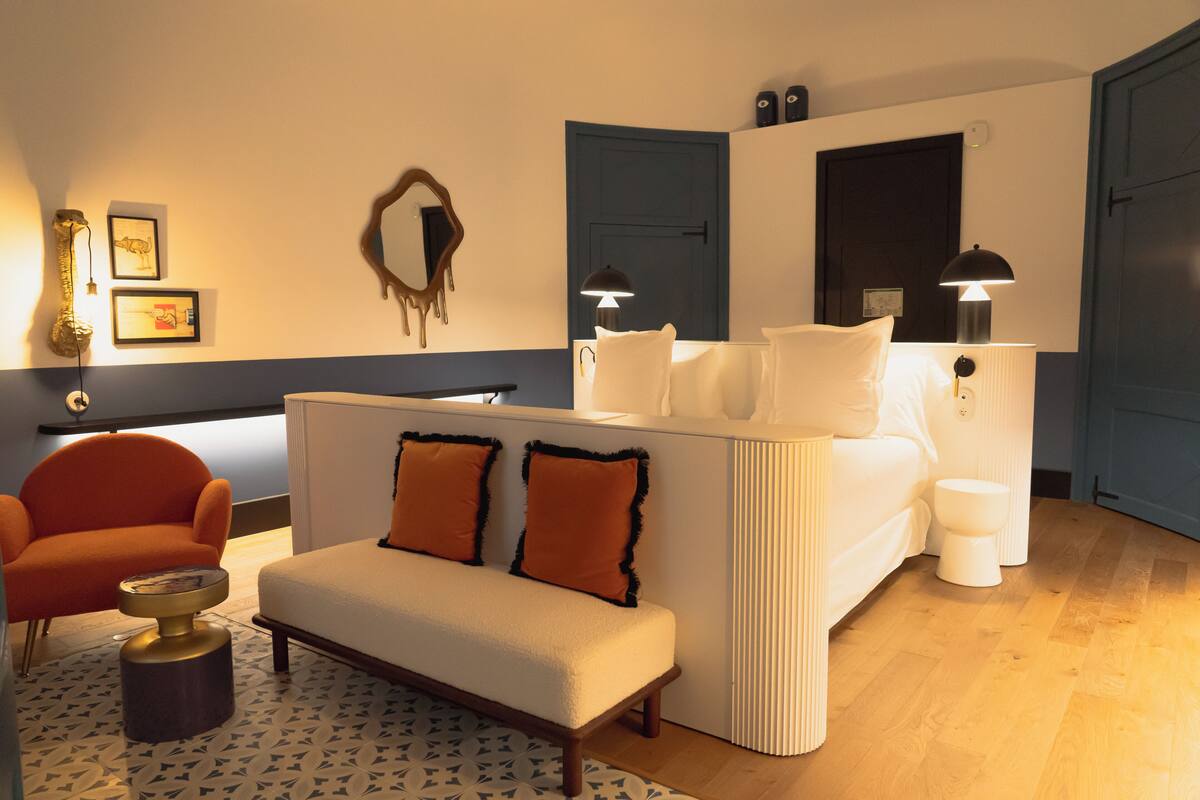 A modern hotel room with a sofa, armchair, bedside table, and a large mirror.