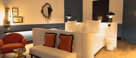 Una habitación de hotel moderna con sofá, sillón, mesita de noche y un espejo grande.