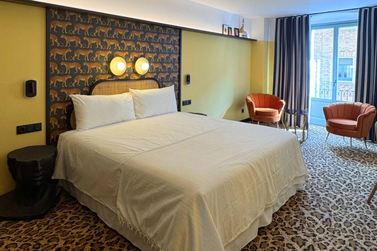 A hotel room with a large bed, two bedside lamps, a leopard print carpet, a chair, and a view of the outside.