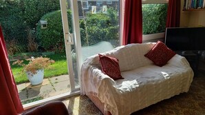 Een kamer met een witte bank, rode kussens, een televisie en uitzicht op een tuin via een glazen deur.