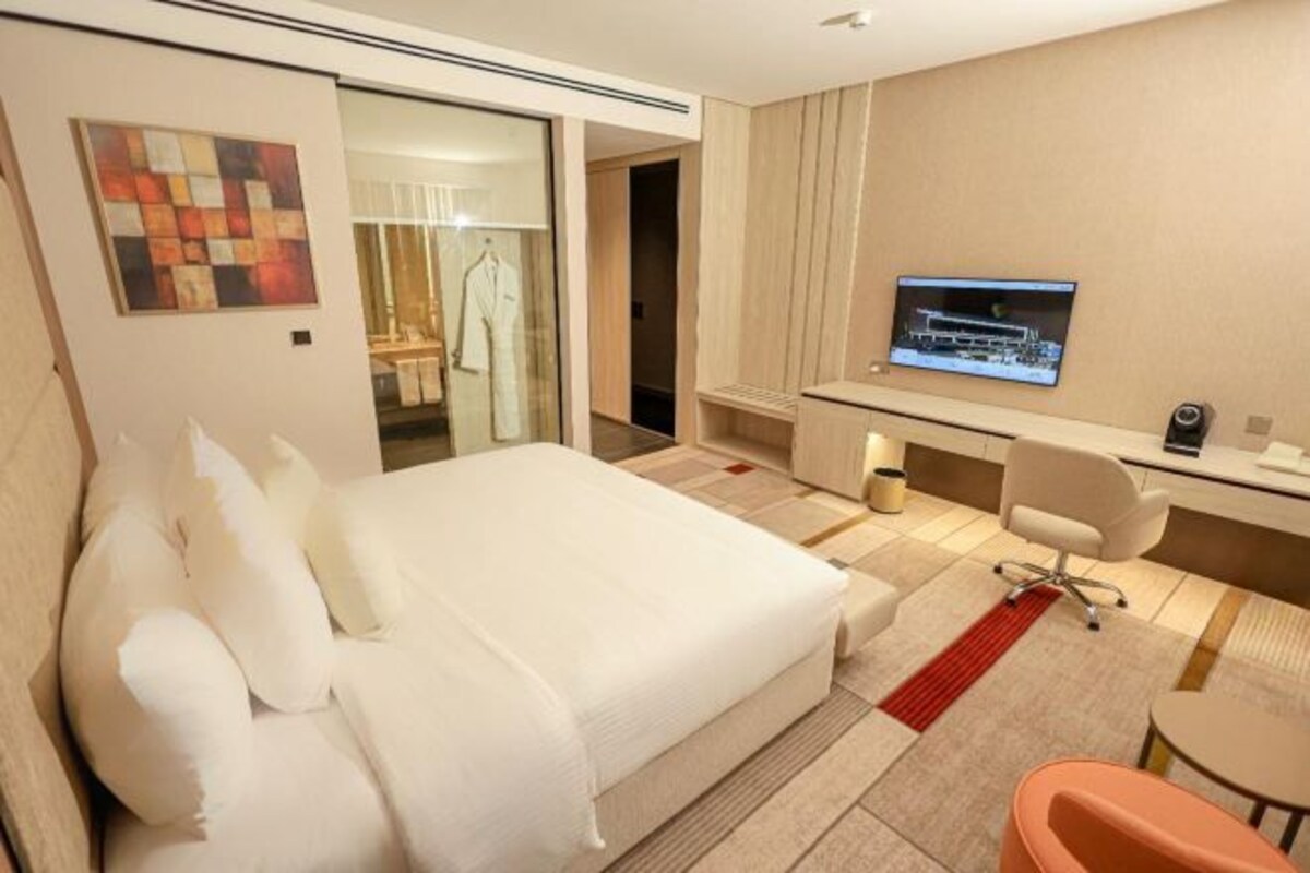 A hotel room with a bed, a desk, a chair, a TV, and a bathroom visible through a glass door.
