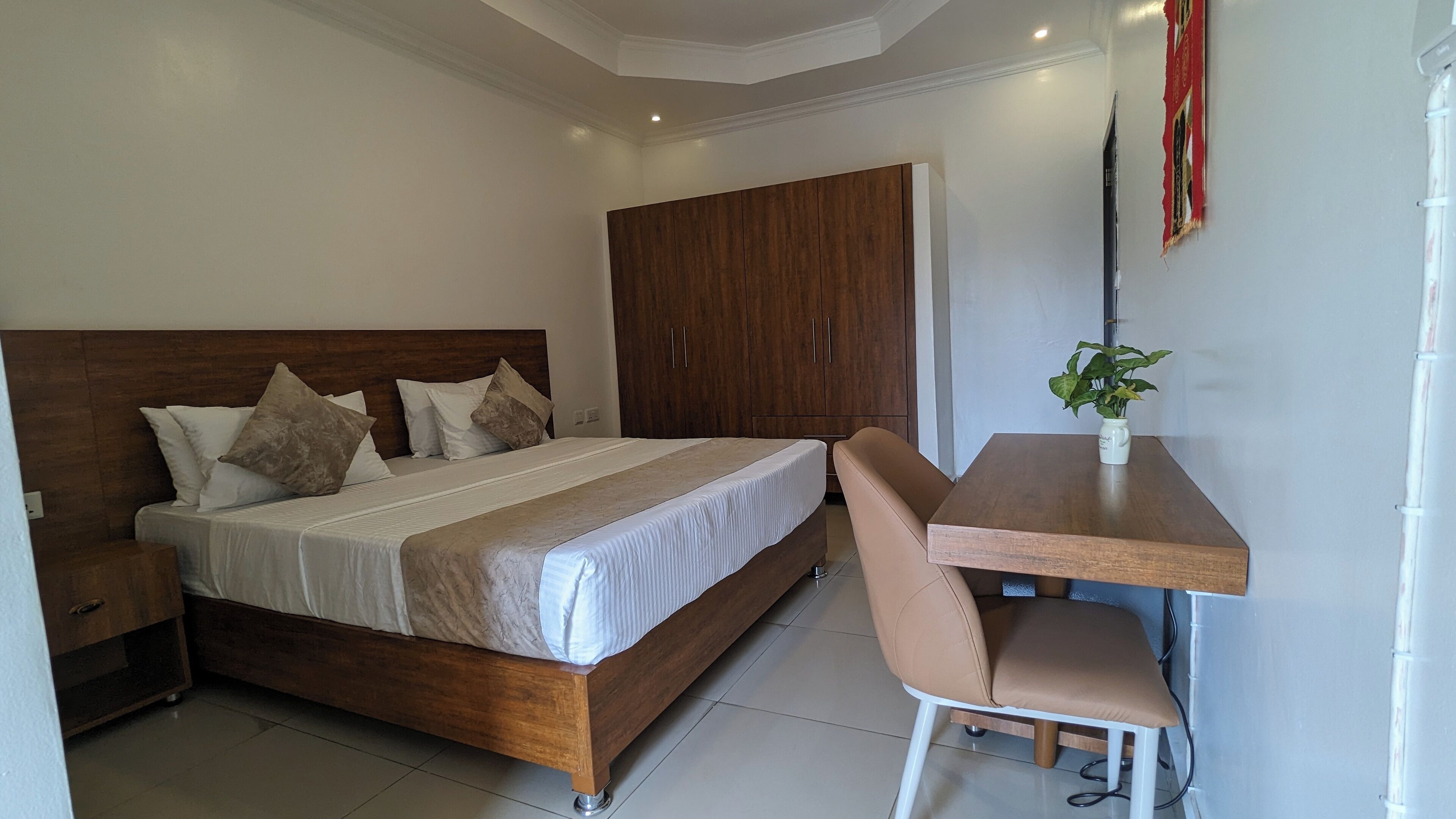 A bedroom with a wooden bed, a wooden wardrobe, a small table with a chair, and a potted plant.