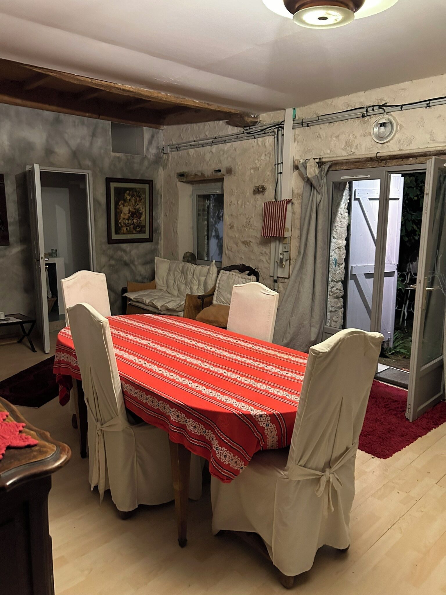 A dining area with a red tablecloth, white chairs, and a view through a glass door.
