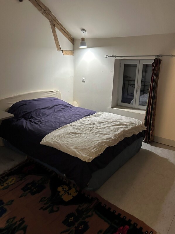 Una cama individual con edredón blanco y azul, cabecero de madera, mesita de noche con lámpara y ventana con cortinas.