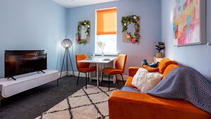 A modern living room with a flat-screen TV, orange furniture, a dining area with a table and chairs, and decorative wall art. - Botanical House Bloom Suite (Blackpool)
