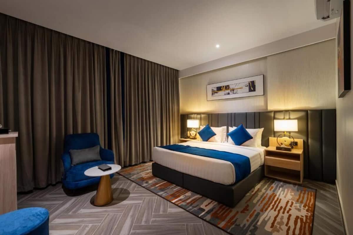 Una habitación de hotel con una cama grande, un sillón azul, una mesita redonda y una mesita de noche con lámparas.