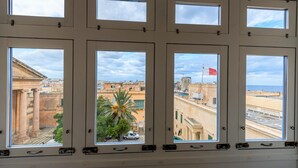 A room with multiple windows offering views of a cityscape, including historical buildings and a beach. - Luxury 2BR Valletta Apartment - Square Sea View (Valletta)