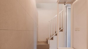 Un pasillo estrecho con una escalera que sube a un piso superior. Las paredes están cubiertas de azulejos y hay un banco a la derecha.