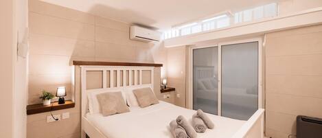 A modern bedroom with a large bed, bedside tables, an air conditioning unit, a window with blinds, and a view of the outside.