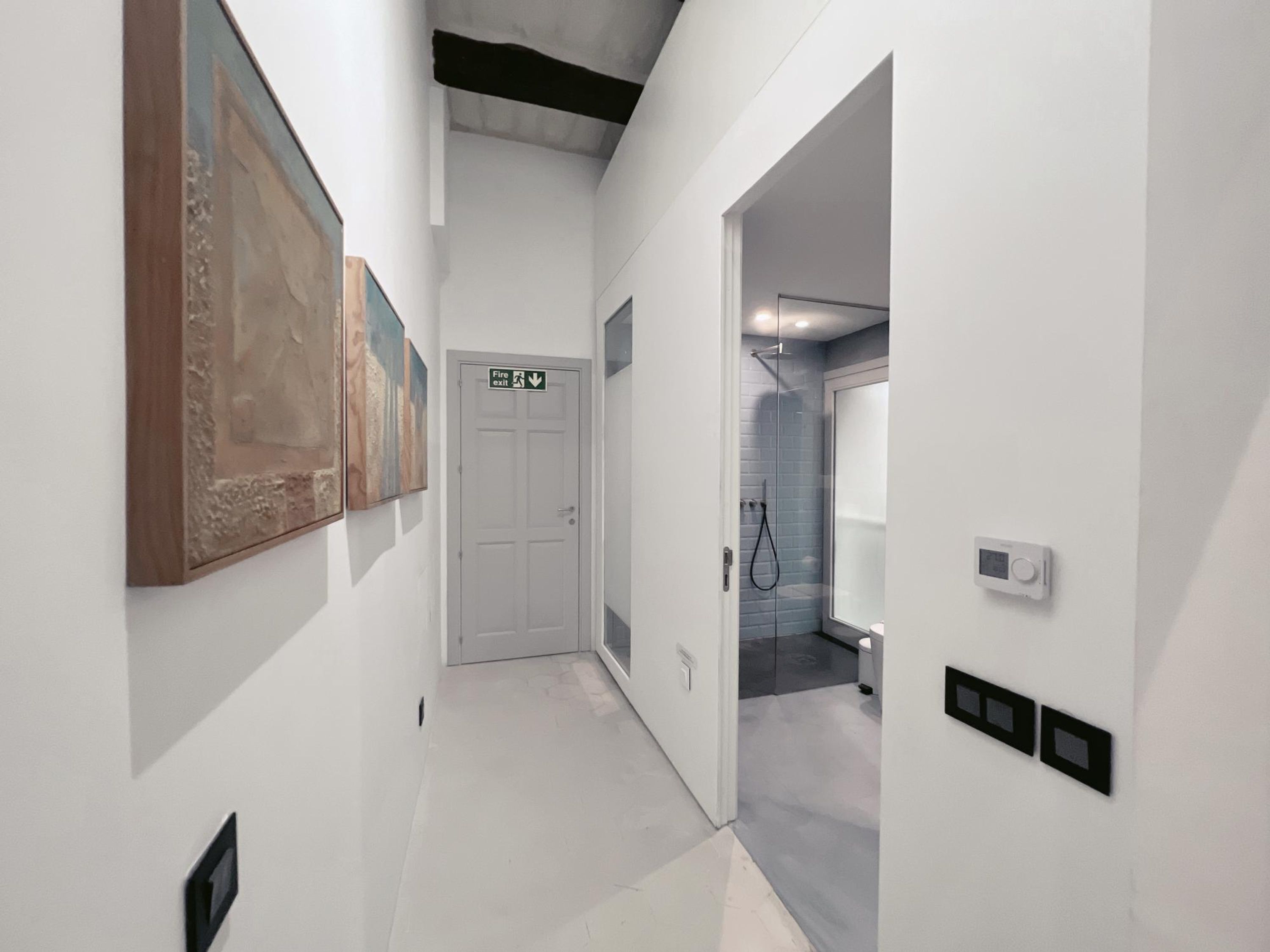 Un couloir aux murs blancs, une porte indiquant « Sortie de secours » et un espace douche doté d’une porte en verre.