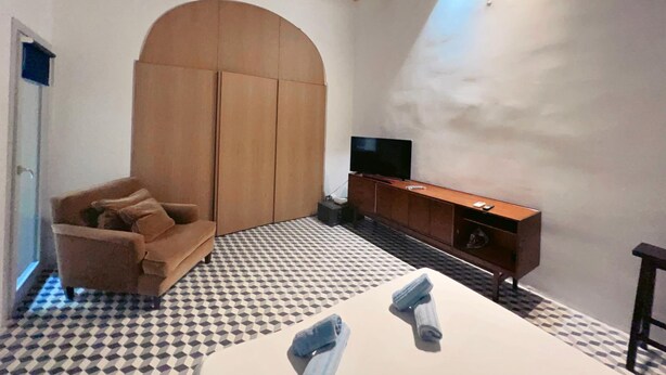 A room with a checkered floor, a sofa, a wooden wardrobe, a TV on a wooden cabinet, and a small table with towels. - Valletta Style 1BR Apartment - Central Location (Valletta)