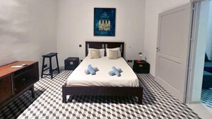 A bedroom with a checkered floor, a bed with pillows, a nightstand, a desk, and a painting on the wall. - Valletta Style 1BR Apartment - Central Location (Valletta)