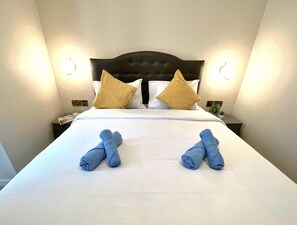 Una cama bien hecha con dos toallas dobladas en el centro, un cabecero oscuro y dos lámparas de noche.