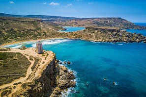 A coastal landscape with a rocky cliff, a small tower, and a beach. - Local Gem Spacious 2BR 2BA Duplex Apartment (Valletta)