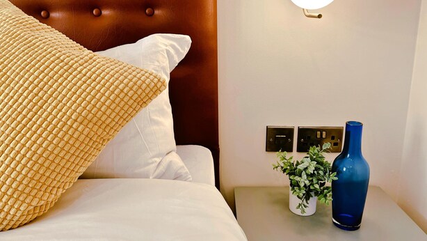 A neatly made bed with a brown headboard, white pillows, and a textured throw. A bedside table holds a small plant and a blue vase. - Local Gem Spacious 2BR 2BA Duplex Apartment (Valletta)