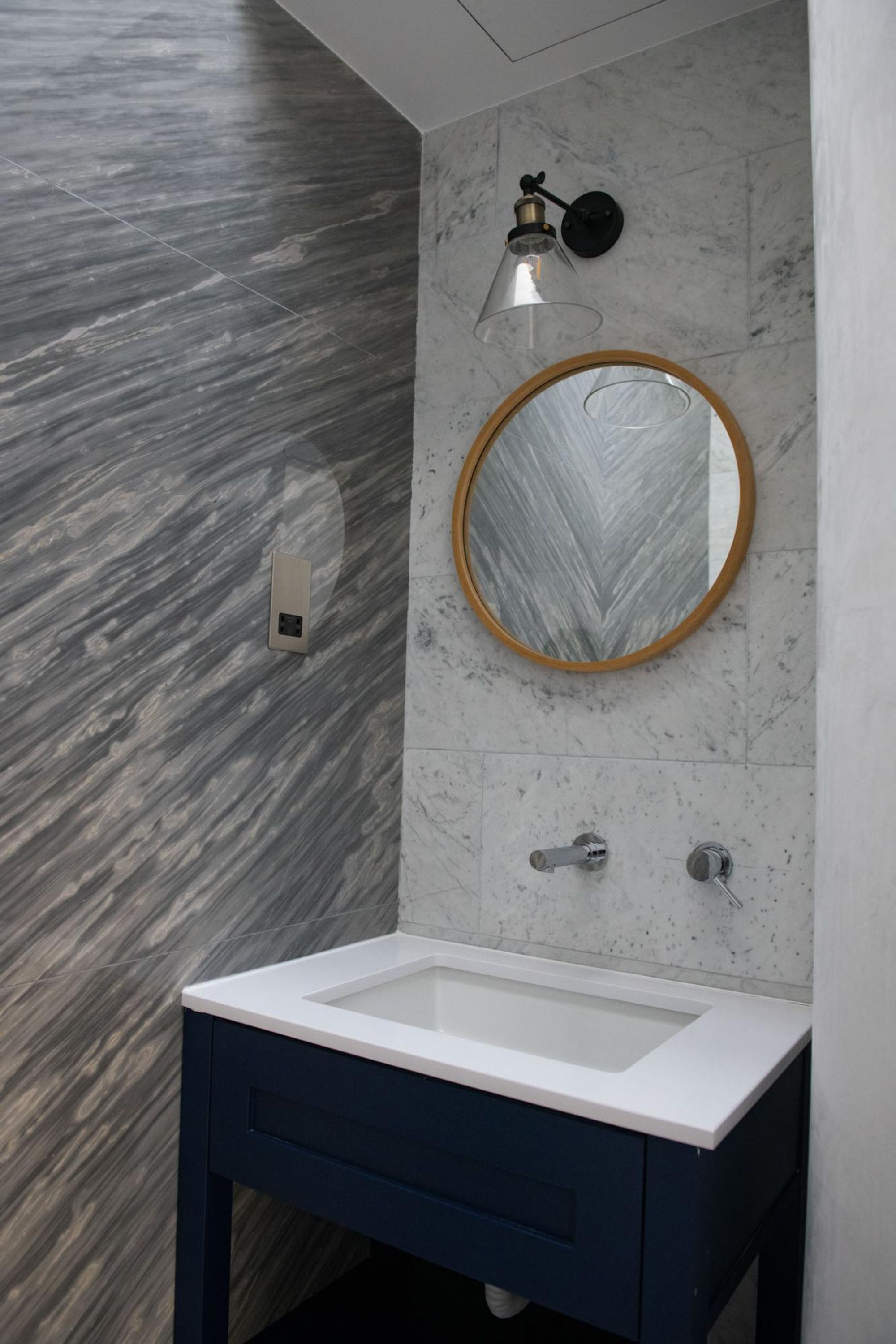 A bathroom with a white sink, dark blue cabinet, marble walls, a round mirror, and a wall-mounted light fixture.