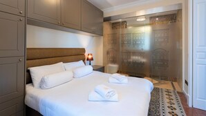A hotel room with a large bed, a headboard, a nightstand with a lamp, and a glass partition with building outlines. - Square and Sea Views - 1 BR With Ensuite (Valletta)