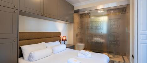 A hotel room with a large bed, a headboard, a nightstand with a lamp, and a glass partition with building outlines.