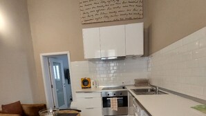 Cocina compacta con muebles blancos, horno empotrado, pileta y heladera pequeña. Hay una mesa de comedor con sillas y un bol sobre ella.