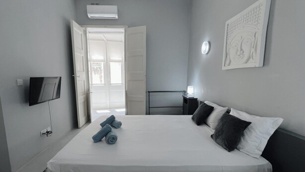 Un dormitorio con cama, televisión, mesita de noche con lámpara, puerta que da a un balcón y una obra de arte colgada en la pared.