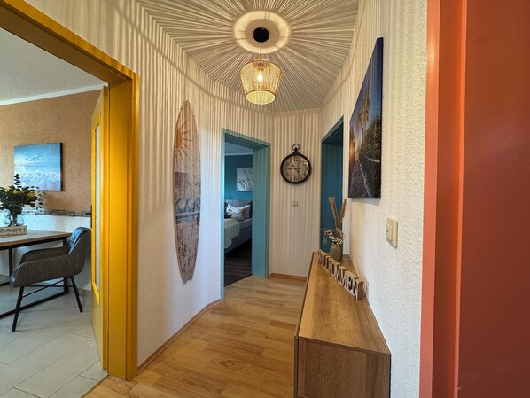 Un pasillo con suelo de madera, techo con lámpara circular y cama con cabecera azul, además de un reloj en la pared.