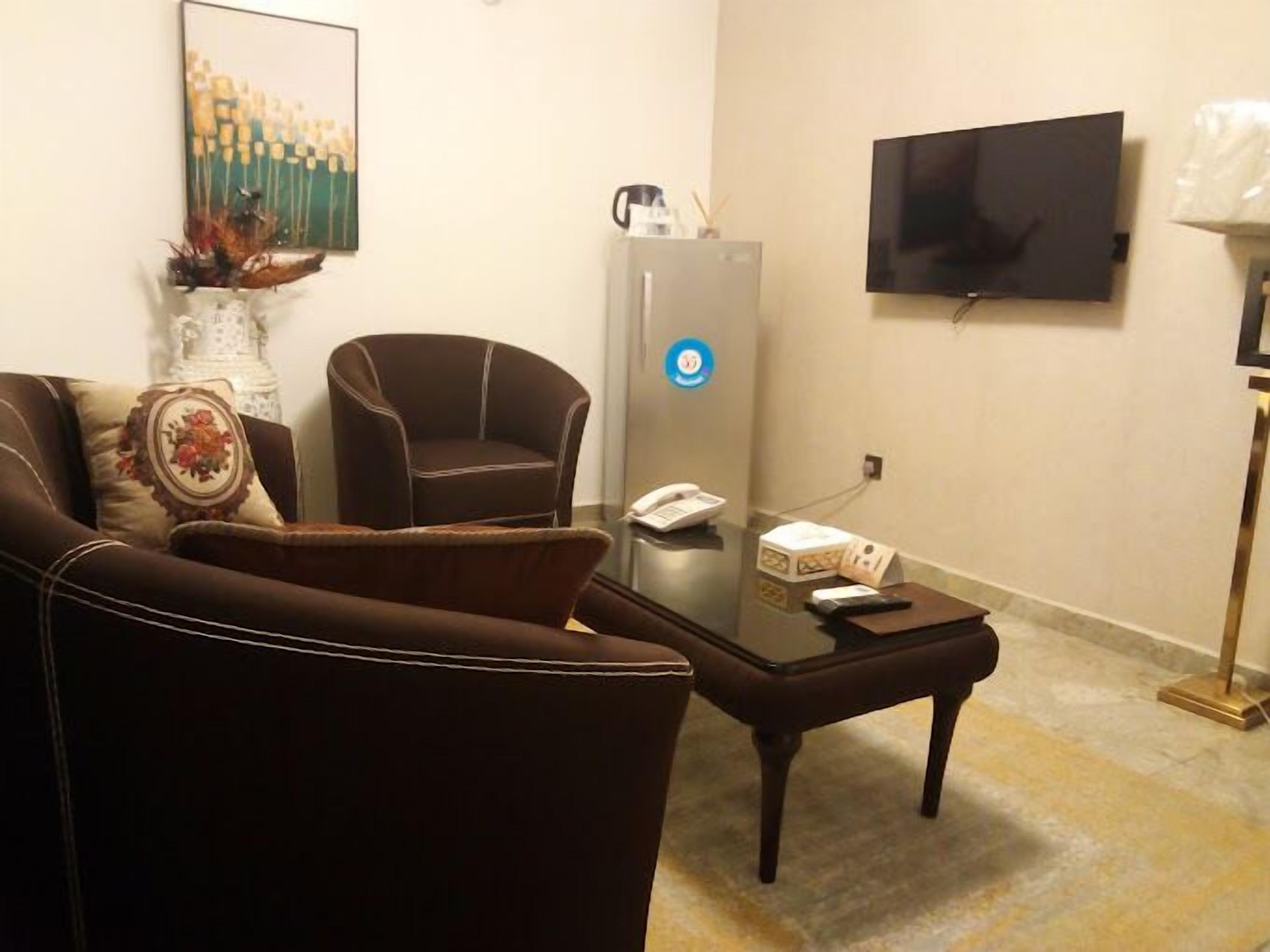 A living room with a brown sofa, a glass coffee table, a white vase with flowers, a brown armchair, a flat-screen TV mounted on the wall, and a refrigerator.