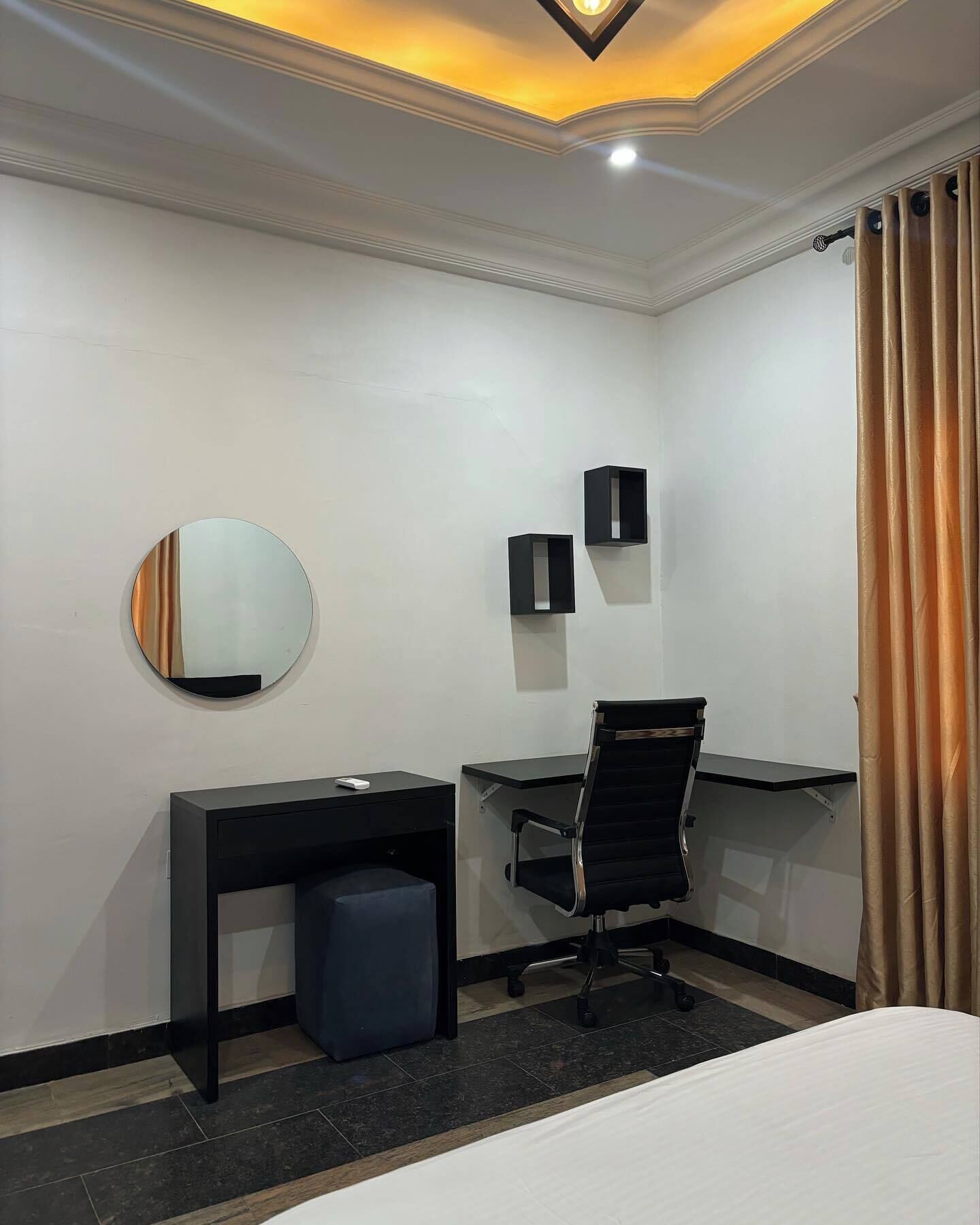 A hotel room with a bed, a desk, a chair, a small stool, a round mirror, and a wall-mounted shelf.