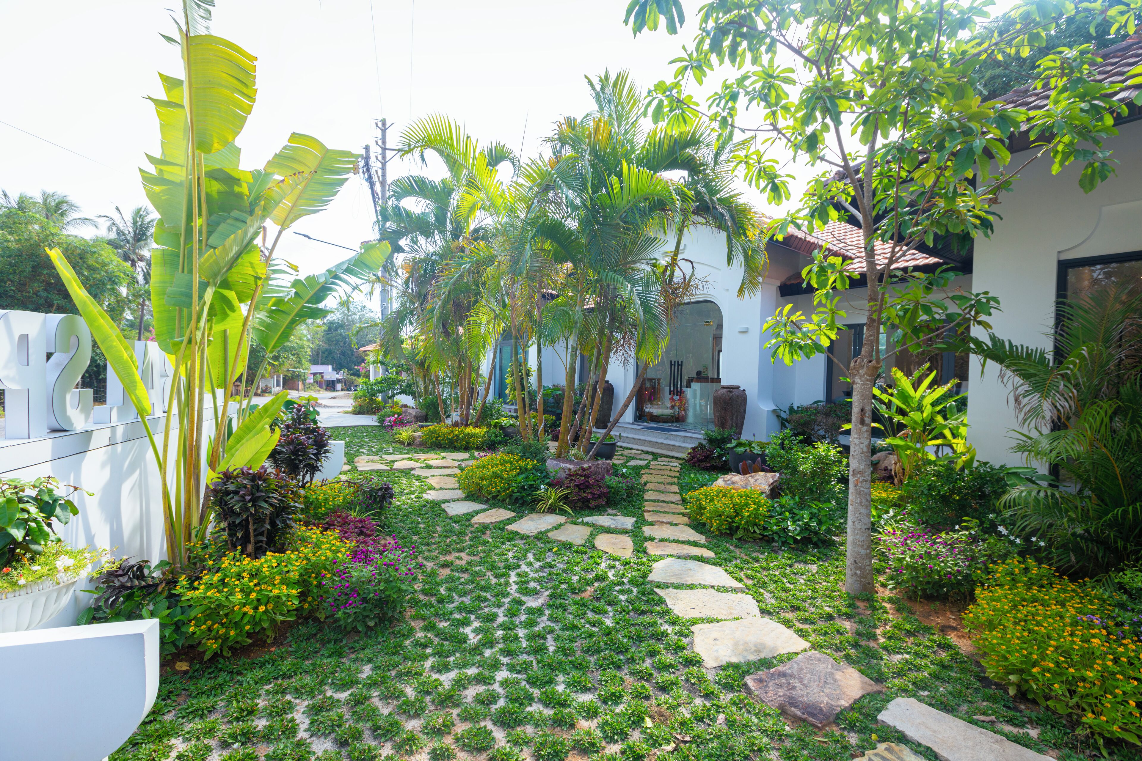 Un sentier de jardin mène à une maison dont le toit est recouvert de tuiles, et qui est entourée de différentes plantes et fleurs.