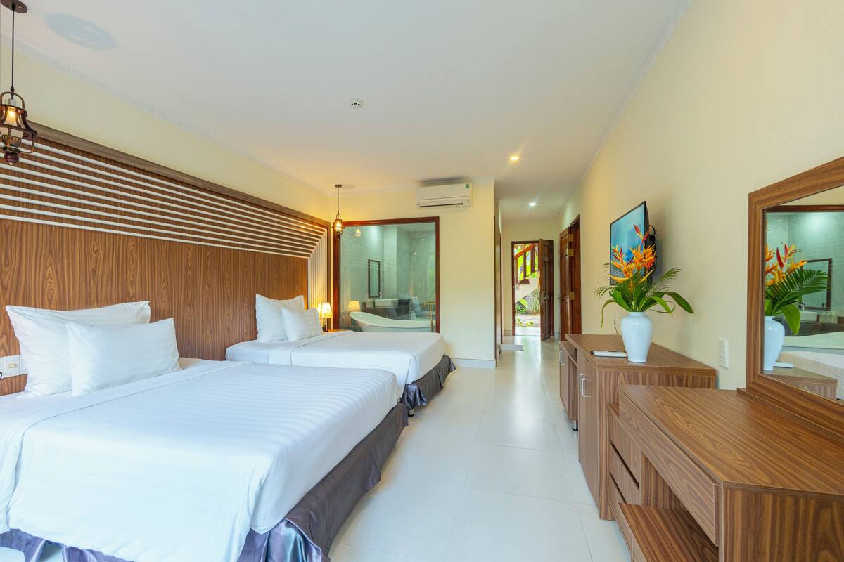 Une chambre d’hôtel avec deux lits, une tête de lit en bois, un grand miroir, une commode en bois et une plante en pot.