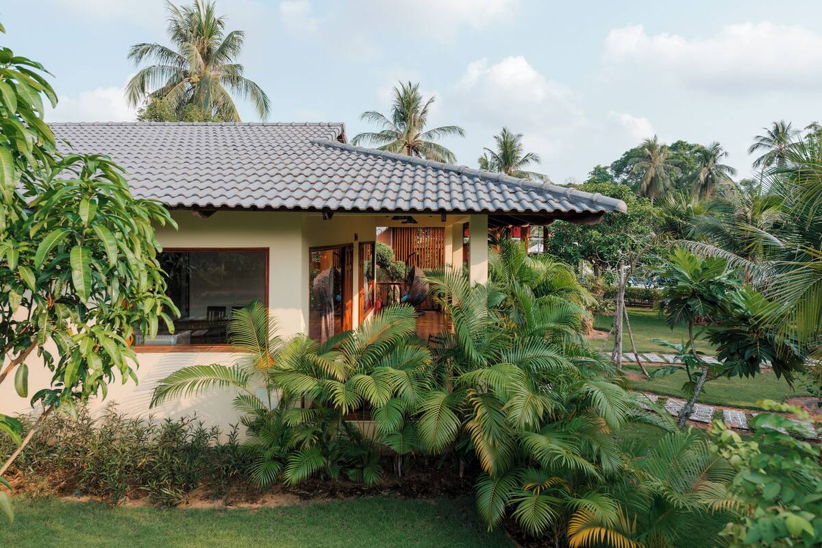 Une maison avec un toit en tuiles, entourée d’une végétation luxuriante et de palmiers.
