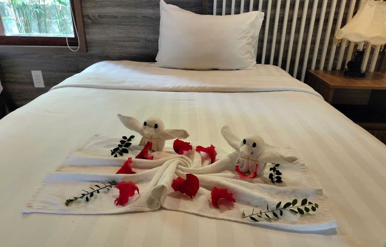 Uma cama bem arrumada com colcha branca, duas toalhas dobradas em formatos de animais e pétalas de rosa vermelhas espalhadas.