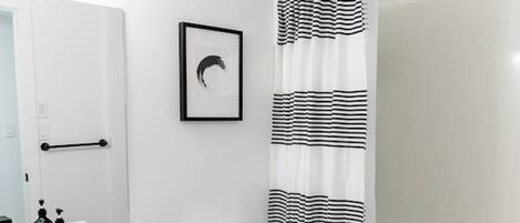 Un baño con inodoro blanco, cortina de ducha a rayas negras y blancas, lavabo blanco con dispensadores de jabón negros y un cuadro en la pared.