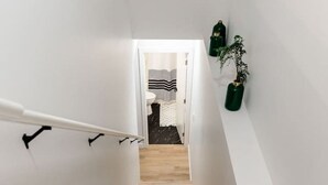 Un escalier étroit avec des marches en bois mène à une pièce dont le mur est blanc et où se trouve une petite plante sur la droite.