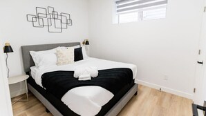 Une chambre à coucher moderne avec une tête de lit grise, du linge de lit blanc et une œuvre d’art murale aux motifs géométriques.
