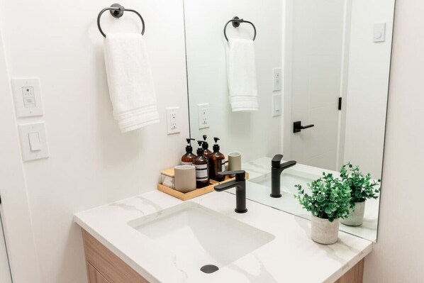 A modern bathroom with a white sink, black faucet, and a mirror reflecting a towel and a door. There are two potted plants on the counter.